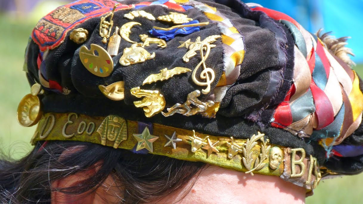 Gros plan sur une faluche portée. Le bandeau doré aligne des étoiles, des palmes et une tête de mort ; le velours au-dessus est couvert d'insignes de métal.