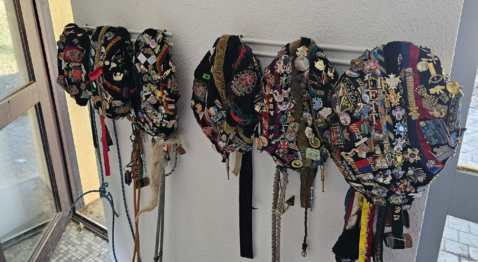 Plusieurs faluches accrochées côte à côte à une barre le long d'un mur. Chacune est couverte d'insignes et de rubans qui pendent jusqu'en bas.