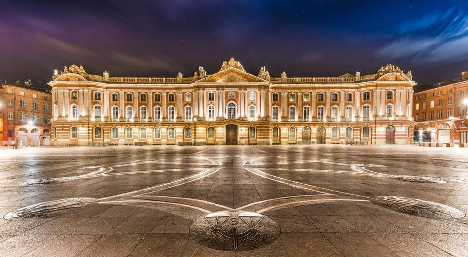 La place du Capitole la nuit, illuminée. La longue façade de brique et de pierre occupe toute la largeur, et le pavé porte un grand décor de bronze.